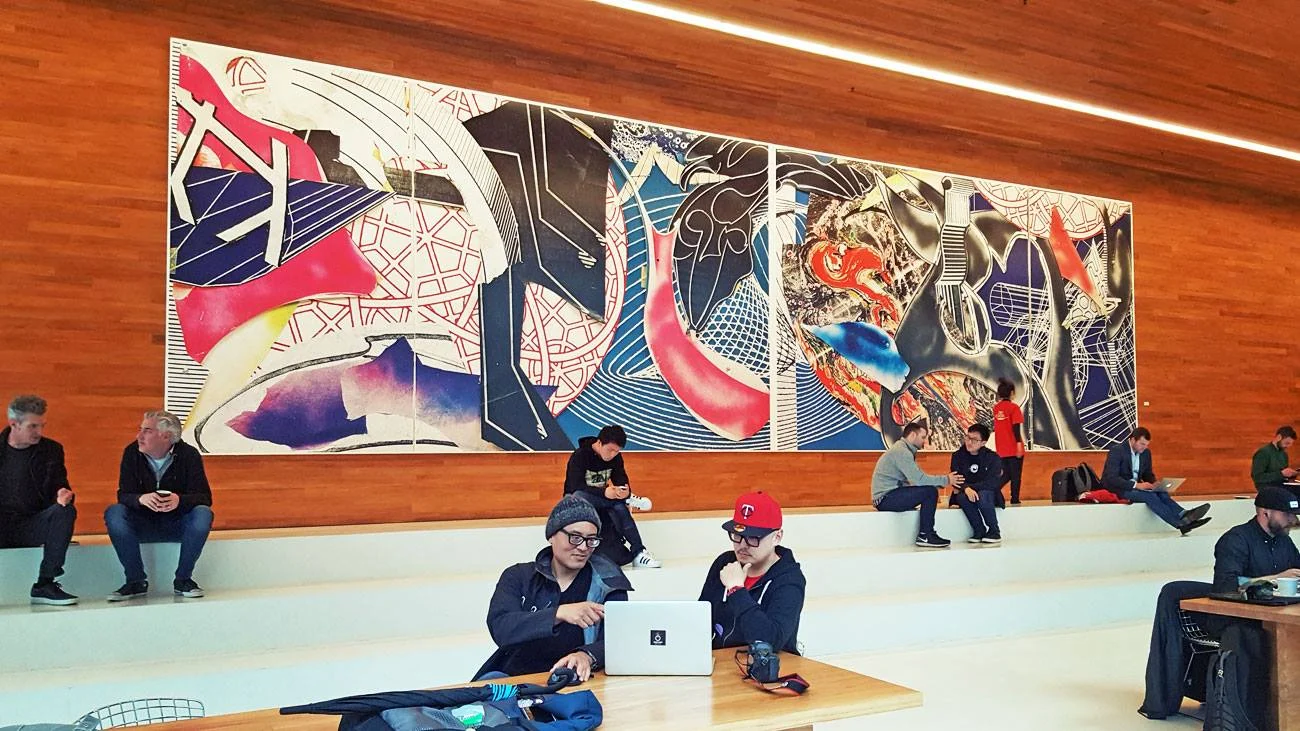 Two people sit at a wooden table working on a silver laptop, while others rest on white steps beneath a large colorful abstract mural with red, blue, and black shapes.