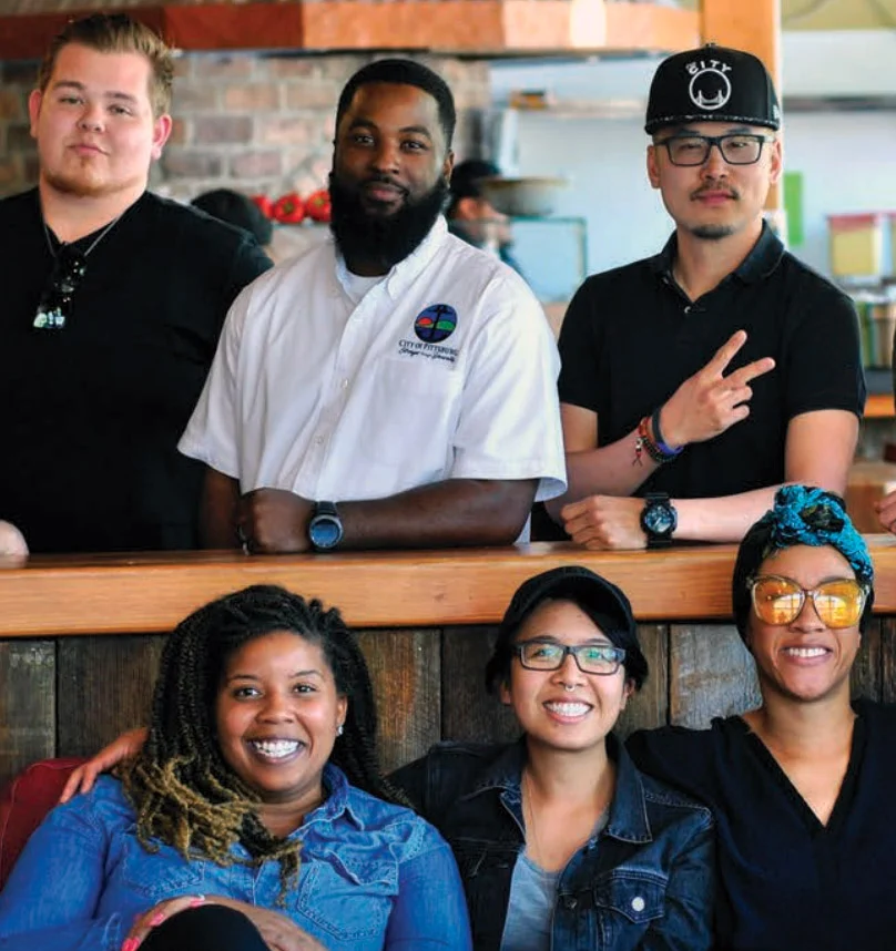 Five people pose at a wooden bar, one in a black cap flashing a peace sign, while three women smile in front.