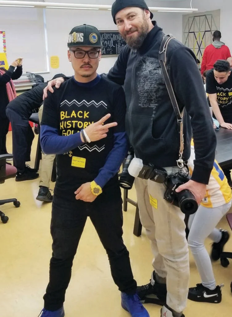 Two men stand close in a bright classroom; the man on the left in a black Black History Month shirt and cap throws a peace sign, while the man on the right in a black jacket with a camera strapped on smiles behind him.