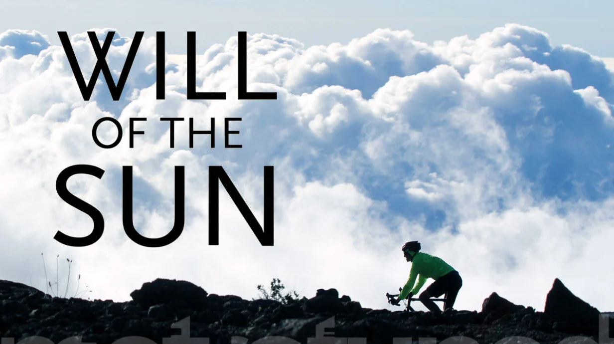 A cyclist in a bright green jacket rides up a dark rocky ridge above a sea of white clouds.