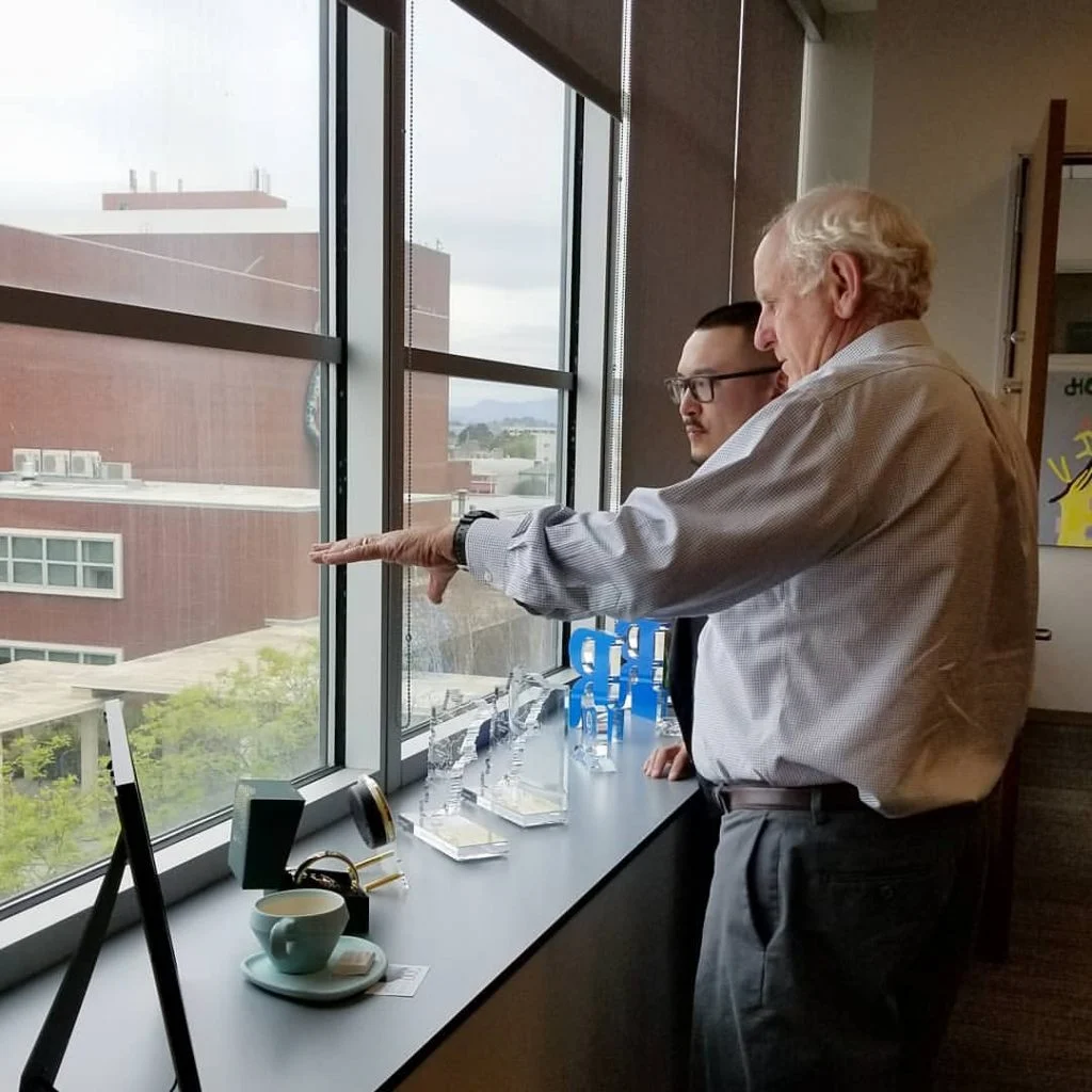 An older man points out a window while a younger man with glasses looks on beside glass awards and a teal cup.