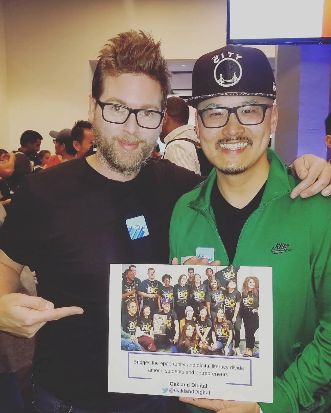 Two men smile indoors, one in a black tee, the other in a green jacket holding a printed group photo.