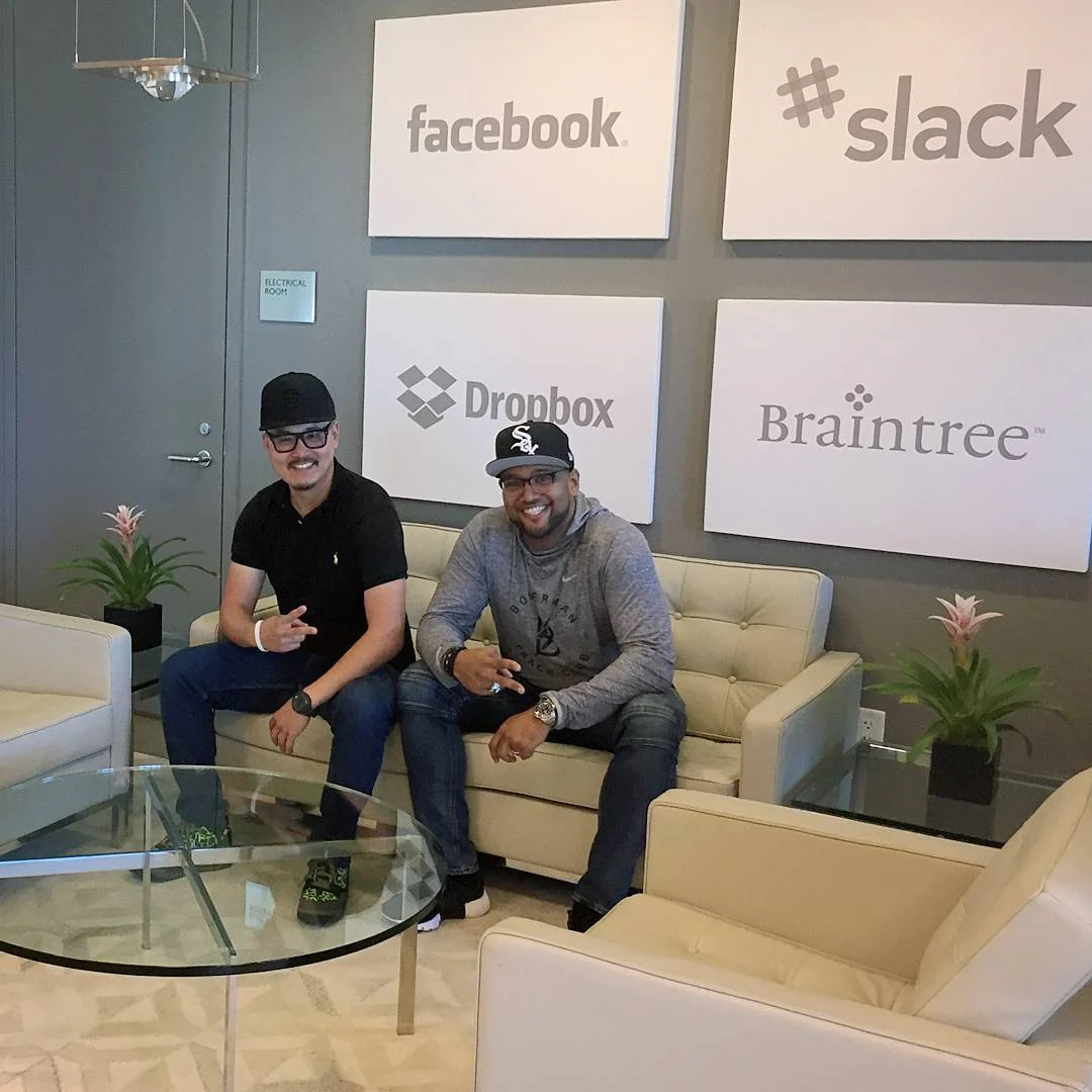 Two men sit smiling on a cream leather couch in an office lobby beneath white logo panels, with a glass table and green plants nearby.