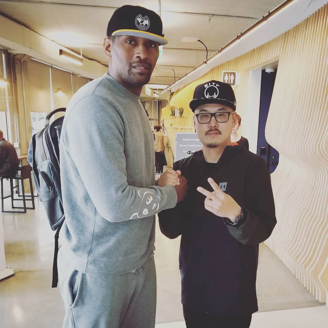 Two men stand indoors in a wood-paneled space, one in a gray sweatsuit and cap clasping hands, the other in a black jacket and cap making a peace sign.