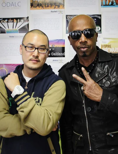 Two men stand indoors before a poster wall, one with crossed arms, the other making a peace sign.