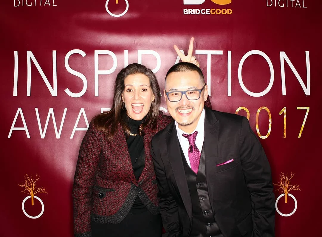 Two people pose in front of a deep red backdrop reading Inspiration Awards 2017, one woman with a bold open-mouthed grin, a man in a black suit and glasses flashing a peace sign behind her.