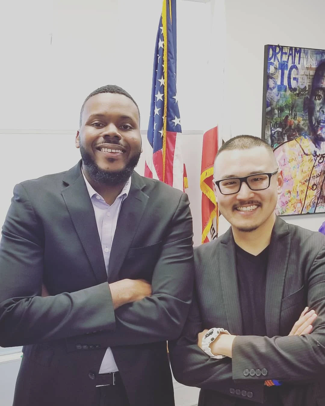 Two men in dark suits stand arm in arm in an office, smiling, with an American flag and a colorful portrait mural behind them.