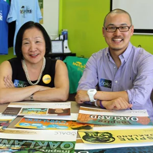 Two people sit smiling at a table covered with colorful printed posters and brochures, with a green wall and hanging shirts behind them.