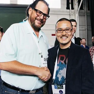 Two men shake hands indoors, one in a light blue shirt and the other in a dark blazer, with a crowd behind.