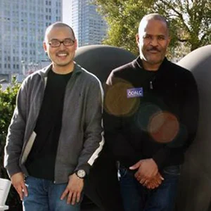 Two men stand outdoors beside a dark rounded sculpture, with city towers and green trees behind them in warm sunlight.