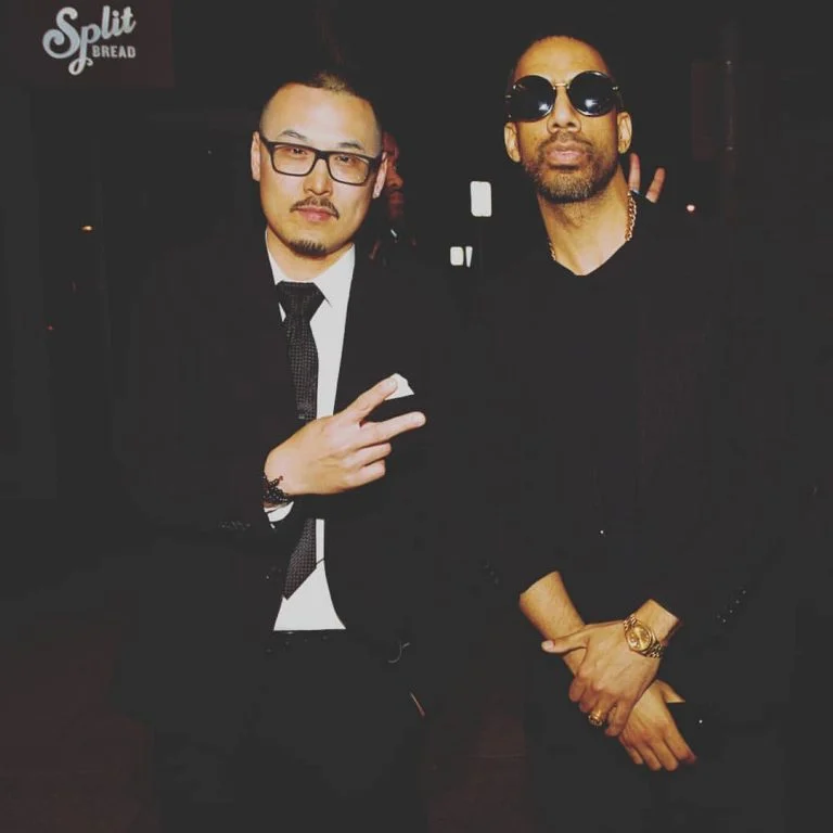 Two men stand in a dark room, one in a black suit flashing a peace sign, the other in black sunglasses and gold jewelry.