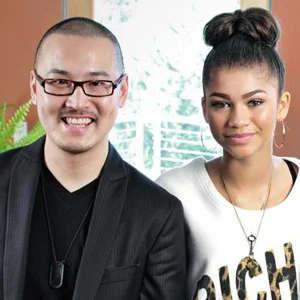 A smiling man in glasses and a dark suit stands beside a woman with a bun before a warm orange wall.