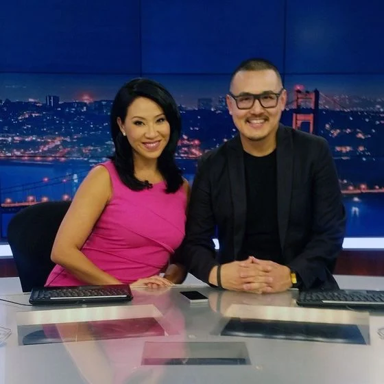Two people sit smiling at a glass news desk in a studio, with a blue skyline backdrop of lit bridges and city lights behind them.
