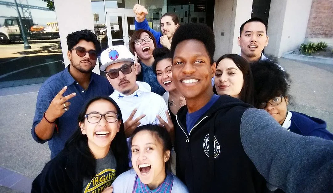 A group of ten young people crowds together outdoors taking a selfie, several making peace signs and raised fists, in front of a glass building with a gray sidewalk and a brick wall.
