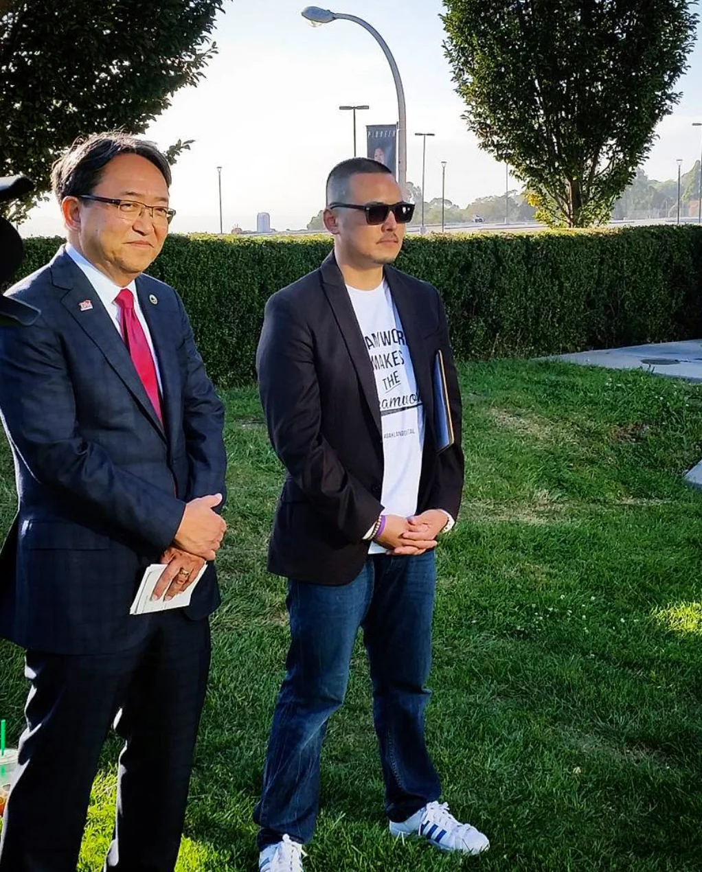 Two men stand on grass before a green hedge, one in a navy suit with red tie, the other in a black blazer and sunglasses.