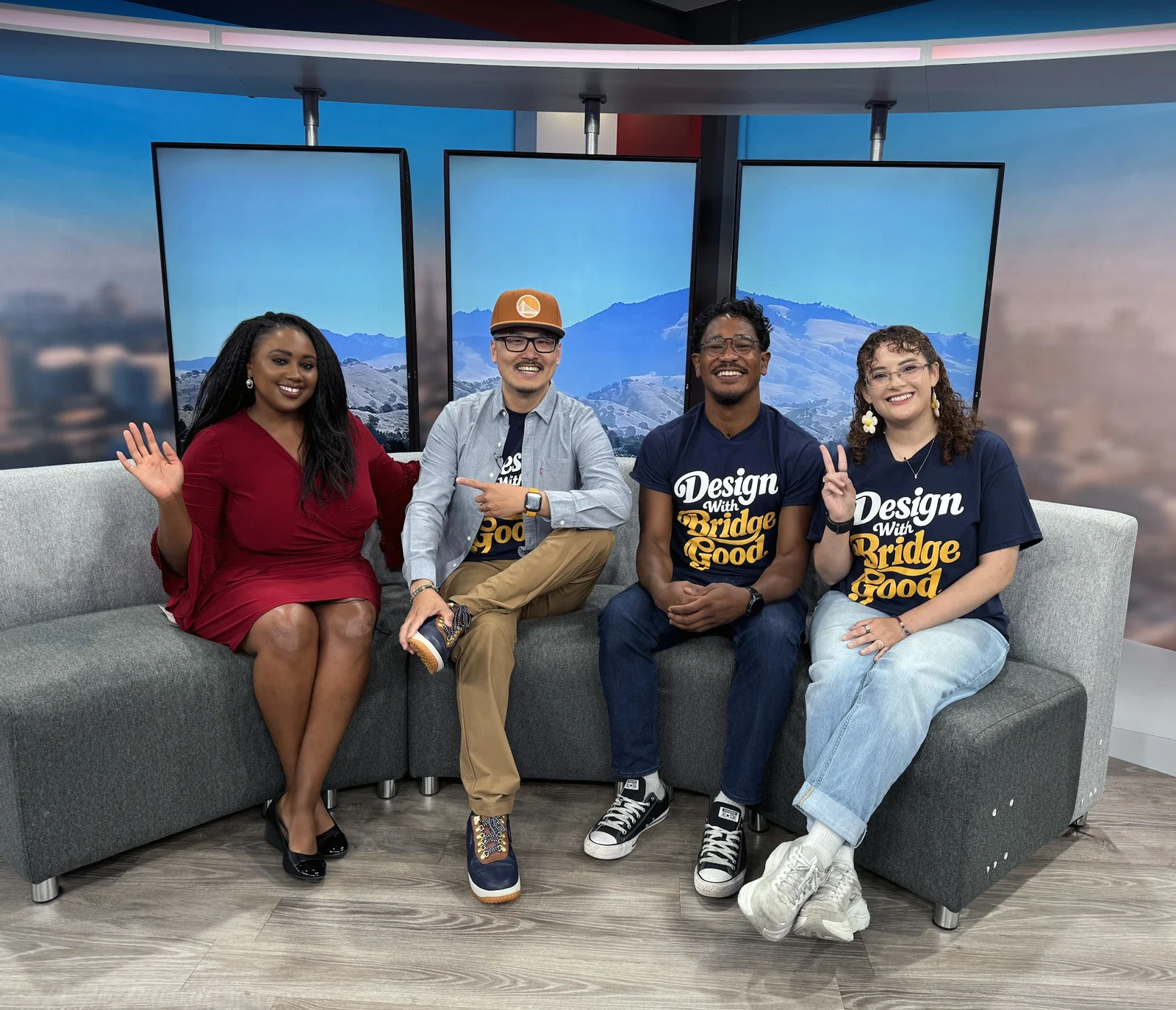 Four people sit smiling on a gray couch in a television studio, one waving, one pointing, two in navy shirts.