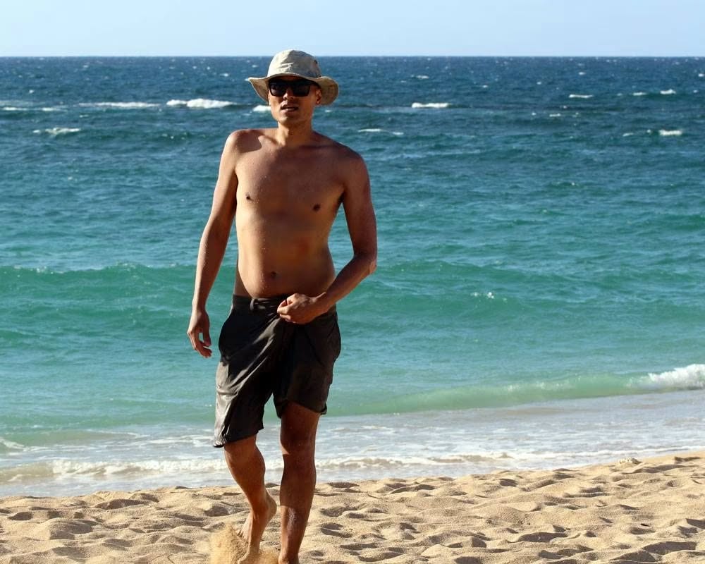 Wilson Tai in a bucket hat and sunglasses walking out of the surf onto a sandy beach.