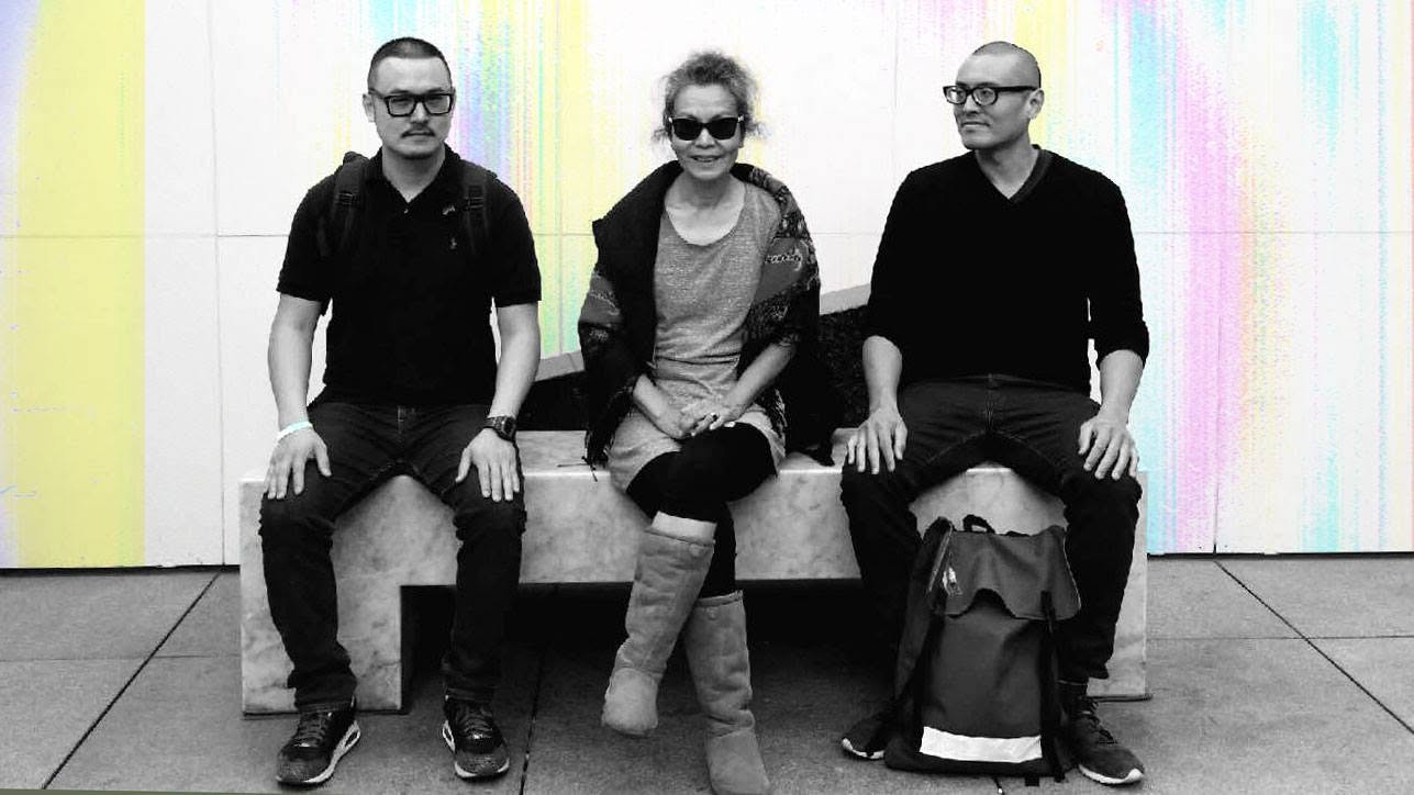 Black-and-white photo of three people seated on a marble bench in front of a pastel-streaked wall.