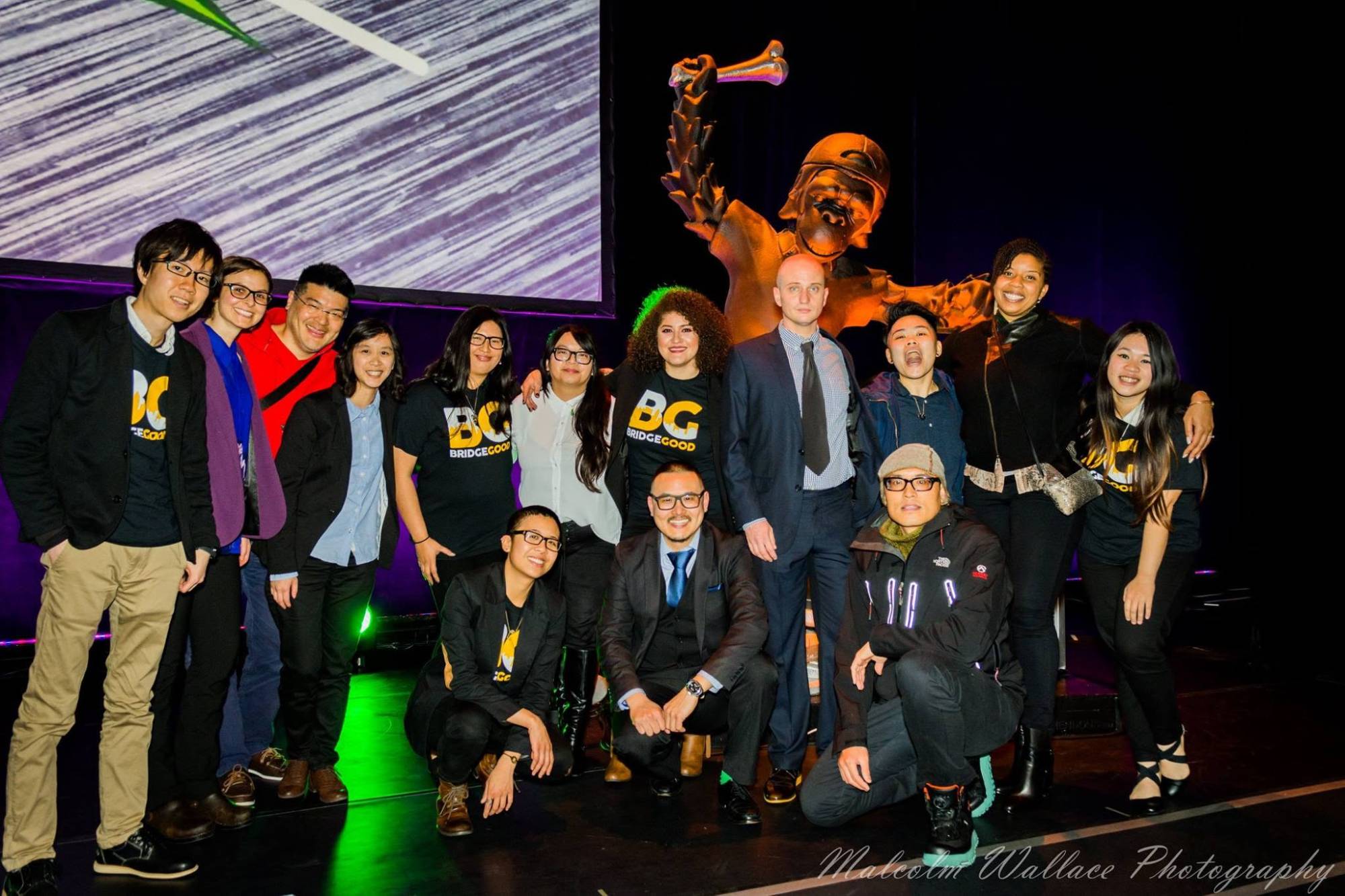Friends and colleagues, some in BridgeGood shirts, pose on a stage in front of a glowing golden statue.