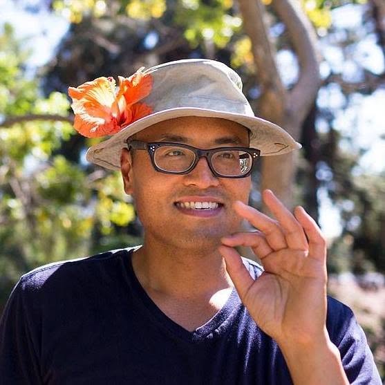 Wilson Tai smiling in a tan bucket hat with an orange hibiscus in the brim, one hand raised.