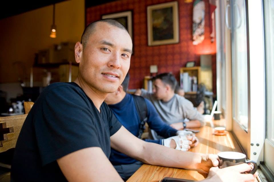 Wilson Tai in a black T-shirt smiling at a wooden café counter in front of a red wallpapered wall.