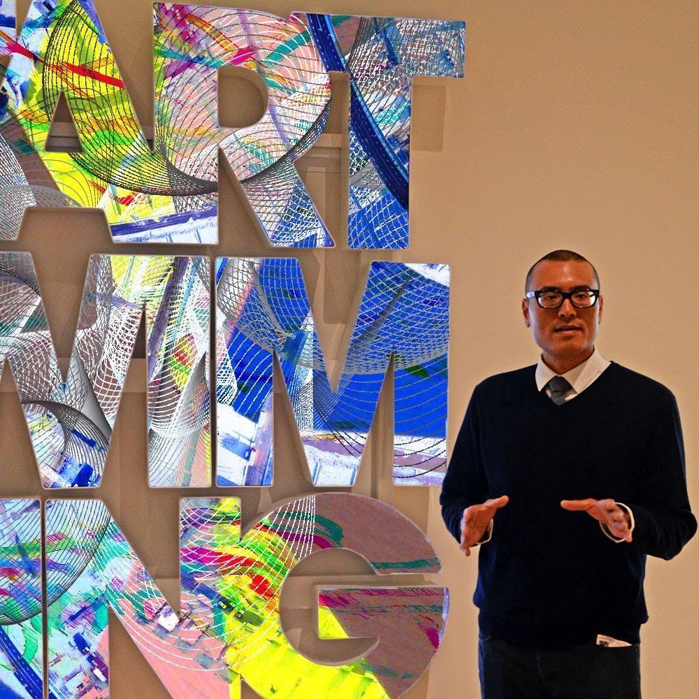 Wilson Tai in black-rimmed glasses and a navy sweater gestures as he speaks in front of large colorful letters.