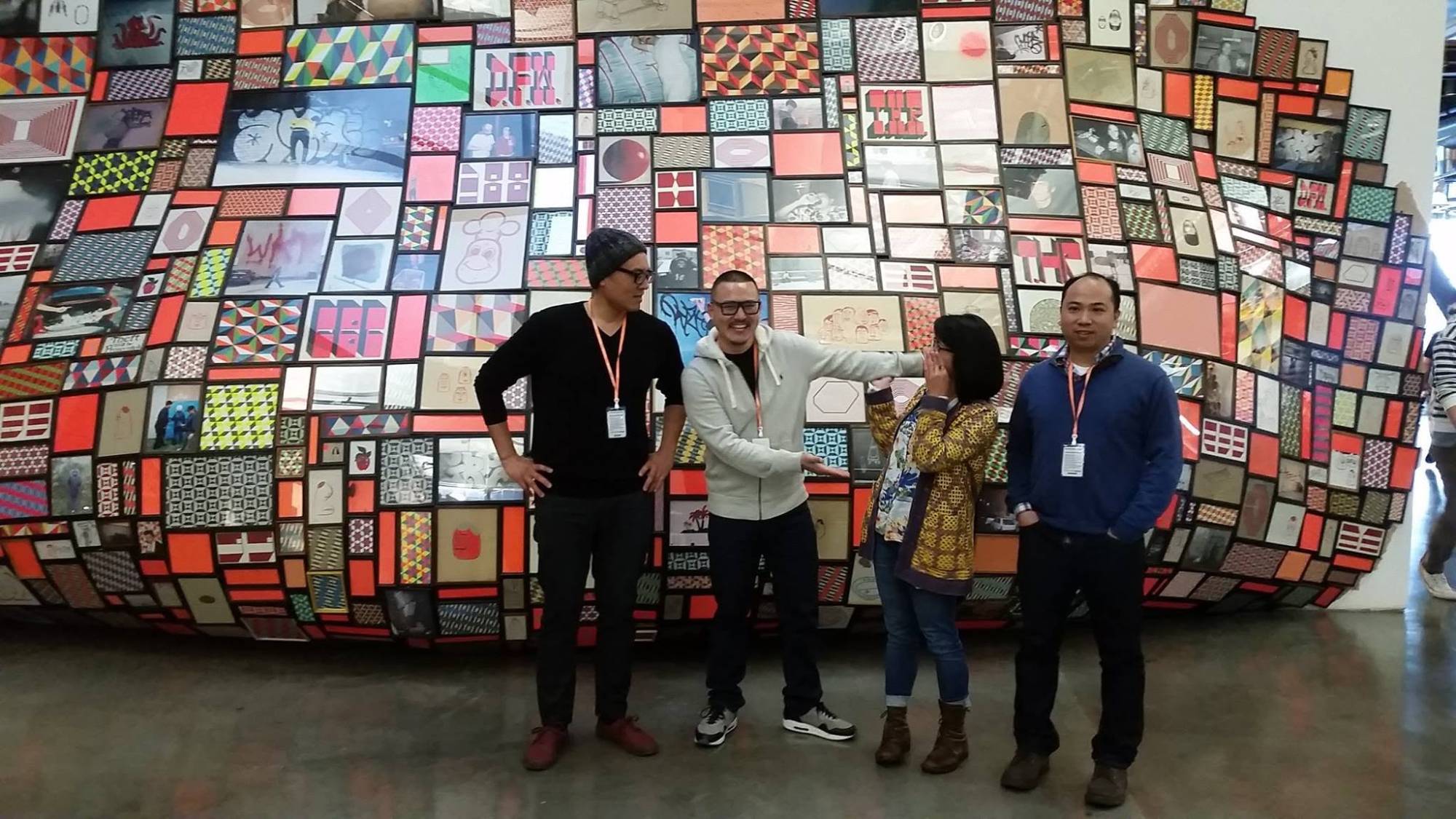 Four people stand in front of a huge wall of colorful framed artwork, two of them joking together.