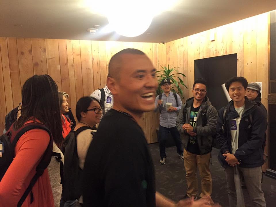 A man laughing mid-turn in a wood-paneled room, with a smiling group in backpacks behind him.