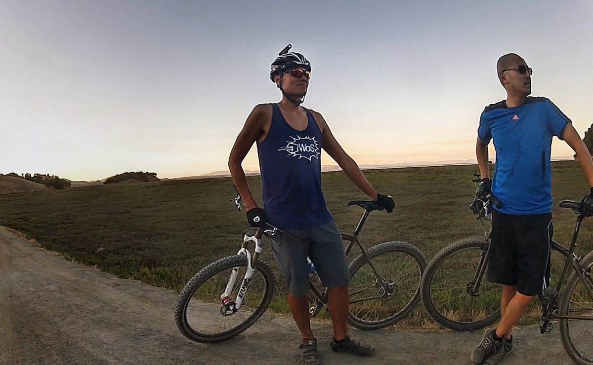 Two cyclists stand with mountain bikes on a dirt road at dusk, one in a blue WoS tank top and helmet.