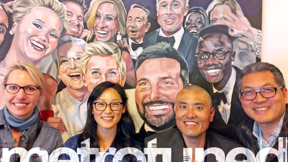 Four smiling people pose in front of a large painted mural of crowded smiling faces.