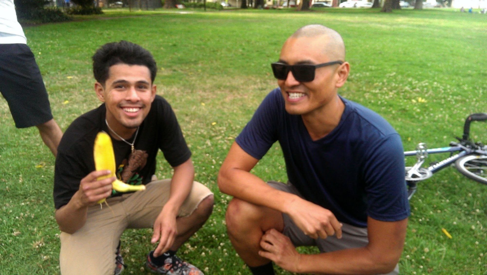 Wilson Tai in sunglasses crouches in a park beside a smiling young man holding a banana.