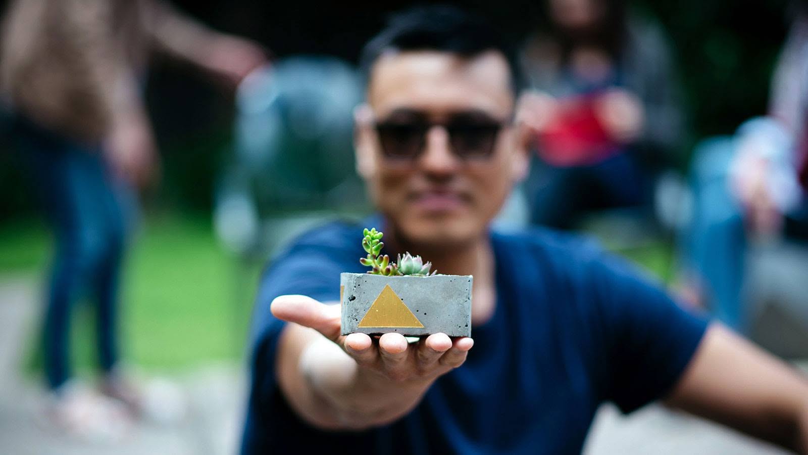 Wilson Tai in sunglasses, blurred behind a hand holding a small concrete planter with a gold triangle.