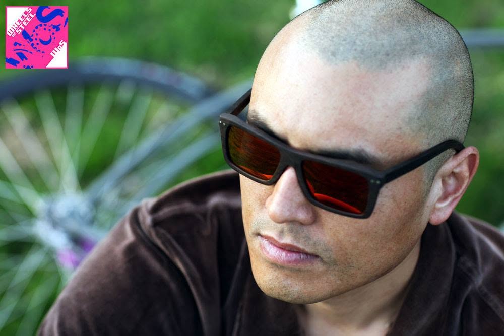 Close portrait of Wilson Tai in dark wooden sunglasses, a bicycle wheel blurred behind him.