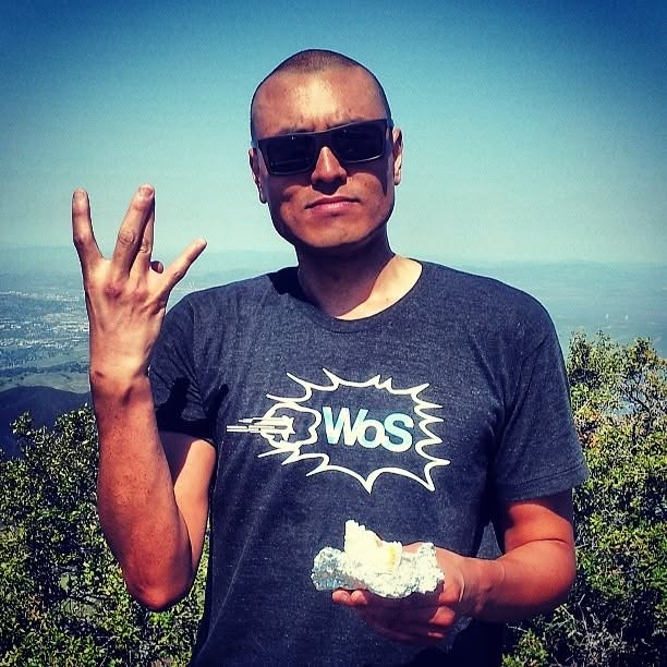Wilson Tai in sunglasses and a WoS T-shirt holding up four fingers on a hilltop above the valley.